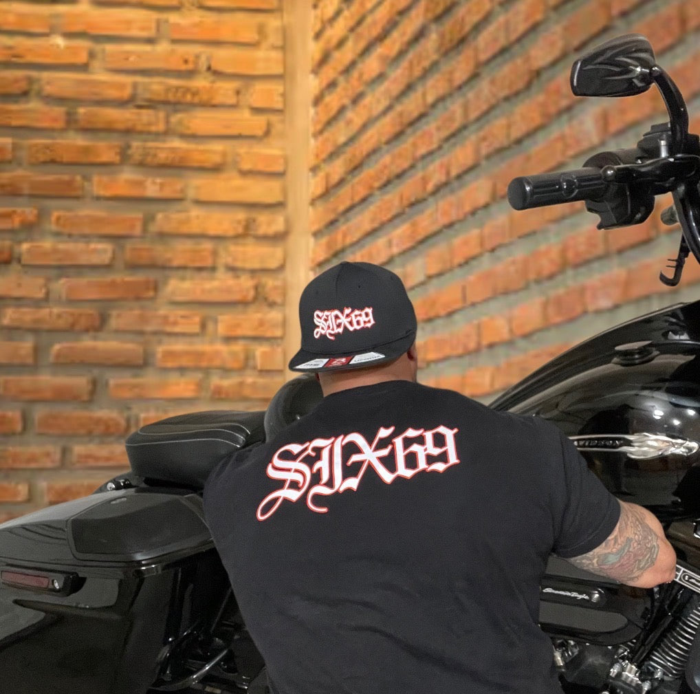 Person wearing a black cap and t-shirt with 'Six69' logo, standing next to a motorcycle against a brick wall.